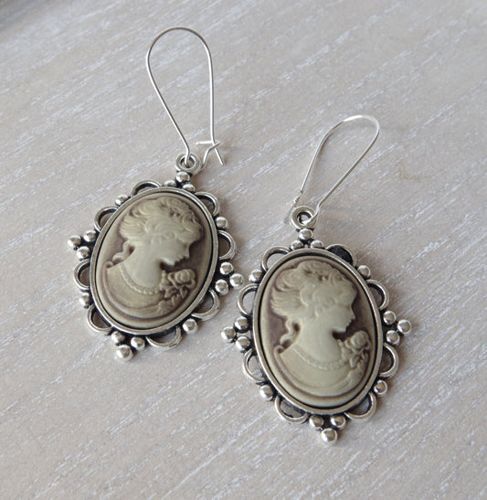 Vue rapprochée de longues boucles d’oreilles au style victorien avec pendentif camée représentant une femme de profil, rehaussé d’un petit nœud décoratif. Teinte bronze patinée, ambiance élégante, fond neutre pour souligner la finesse artisanale du bijou.