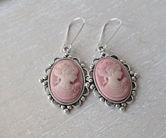 d’oreilles pendantes avec camée rose ovale et profil féminin ivoire, finition bronze antique, posées sur un fond romantique clair. Mise en valeur du style vintage et du raffinement artisanal.